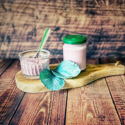 Rotkohl-Pesto mit Pinienkernen in einem Glas und einem kleinen Schälchen auf einem Holzbrett, dekoriert mit frischen Rotkohlblättern.