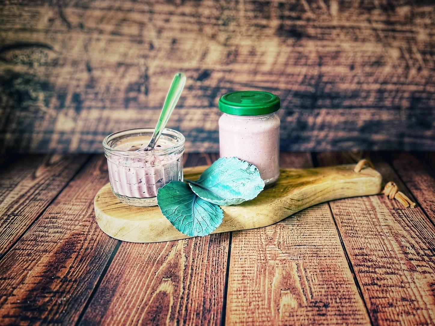 Rotkohl-Pesto mit Pinienkernen in einem Glas und einem kleinen Schälchen auf einem Holzbrett, dekoriert mit frischen Rotkohlblättern.