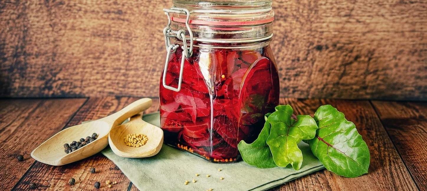 S&uuml;&szlig;-sauer eingelegte Rote Bete in einem verschlossenen B&uuml;gelglas auf einem rustikalen Holztisch, dekoriert mit frischen Rote-Bete-Bl&auml;ttern und Gew&uuml;rzen.