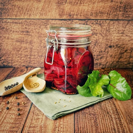 Süß-sauer eingelegte Rote Bete in einem verschlossenen Bügelglas auf einem rustikalen Holztisch, dekoriert mit frischen Rote-Bete-Blättern und Gewürzen.