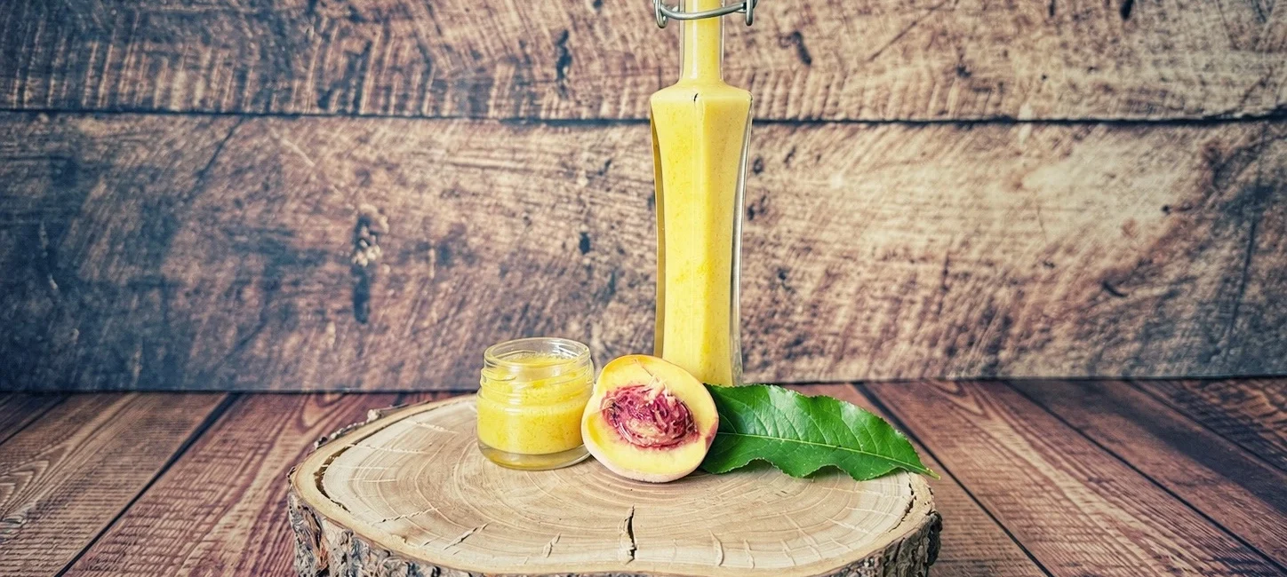 Zwergpfirsich-Dressing in einer B&uuml;gelflasche mit kleinem Glas und halbiertem Pfirsich auf einer Holzscheibe