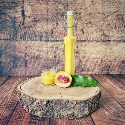 Zwergpfirsich-Dressing in einer Bügelflasche mit kleinem Glas und halbiertem Pfirsich auf einer Holzscheibe
