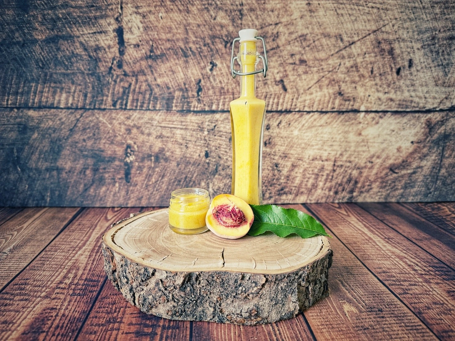 Zwergpfirsich-Dressing in einer Bügelflasche mit kleinem Glas und halbiertem Pfirsich auf einer Holzscheibe